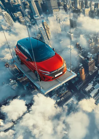 Red and black Nissan vehicle suspended on platform above city skyline and clouds, creating dramatic aerial promotional imagery.