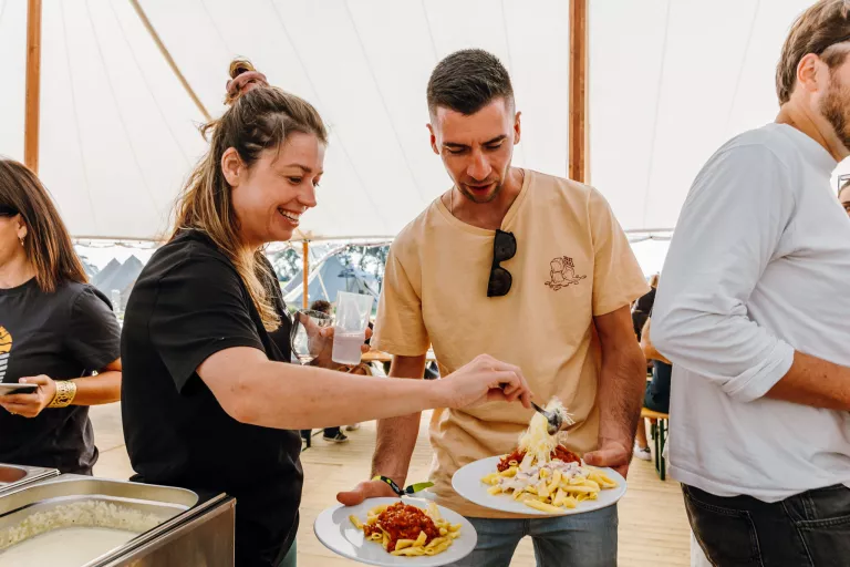 Two people are smiling and serving pasta onto plates from a buffet table under a large white tent at Springbok's summer team event. 