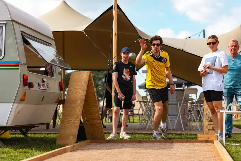 A man wearing a yellow sports jersey plays pétanque outdoors at Springbok's summer team event. Three people watch. A vintage caravan and tents are in the background.