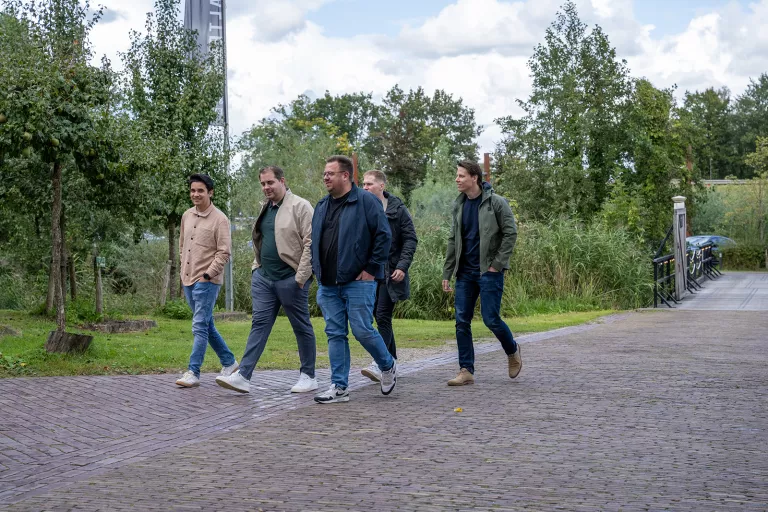 Five men casually walk on a cobblestone path surrounded by lush greenery and trees toward the Dutch Springbok summer team event.