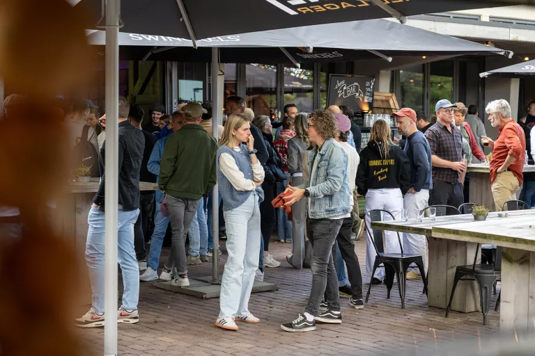 Outdoor social gathering at a bar with diverse group of people under umbrellas. Casual conversation and laughter create a lively and relaxed atmosphere.