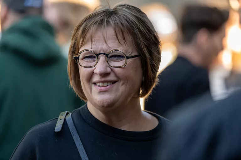 Close-up of a woman with short brown hair and round glasses smiles pleasantly, wearing a black top. Blurred people and warm lighting create a joyful atmosphere.