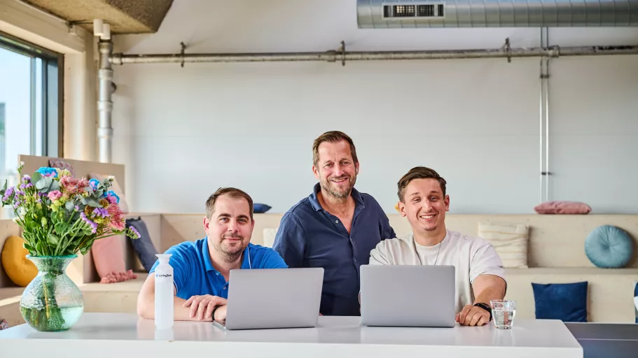 Three Springbok team members sit smiling at a desk with laptops, in a bright office with pastel decor. A vase of colorful flowers adds a cheerful touch.
